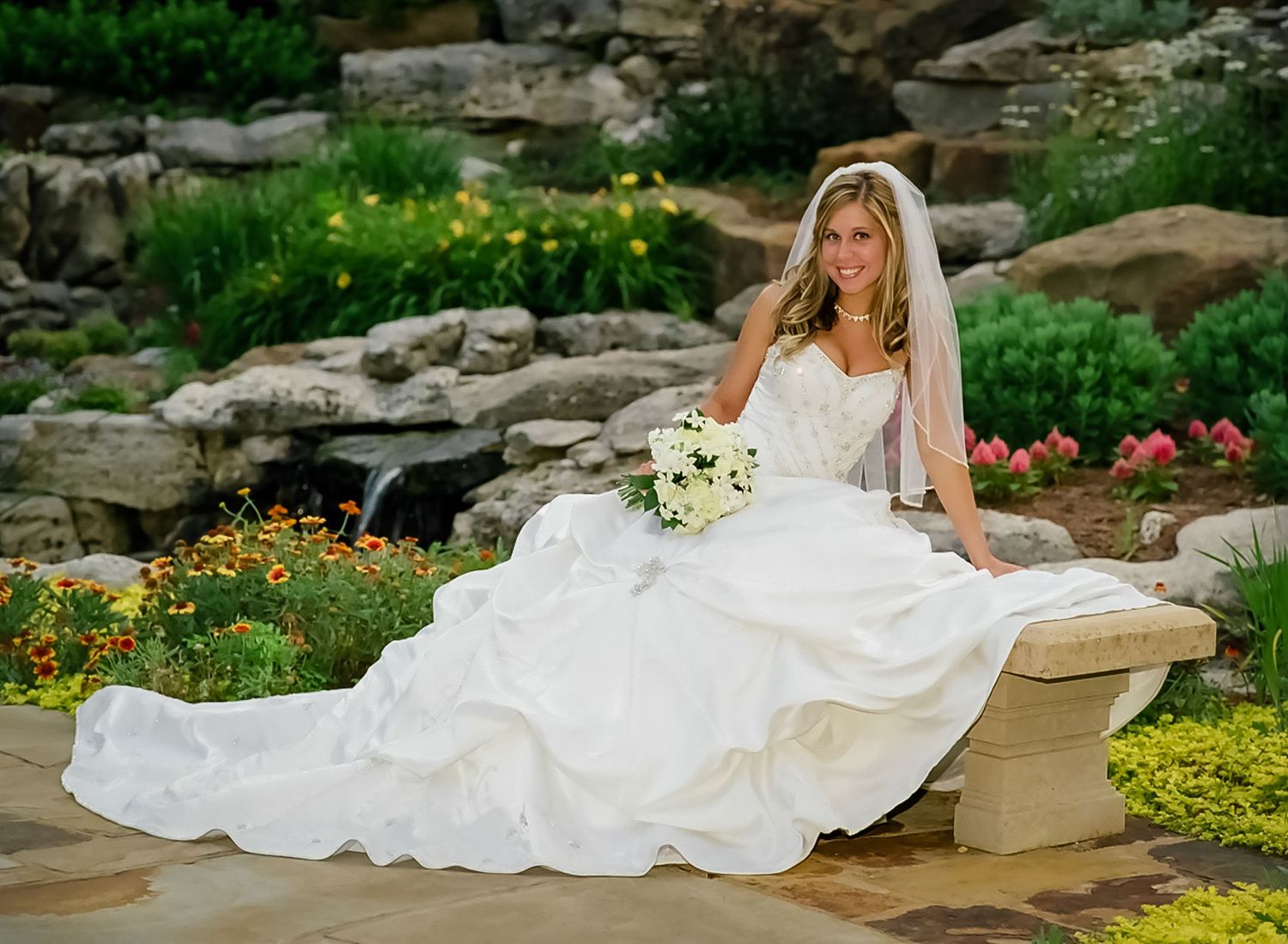Bride in flowing white dress reclines on stone wall amid garden landscape.