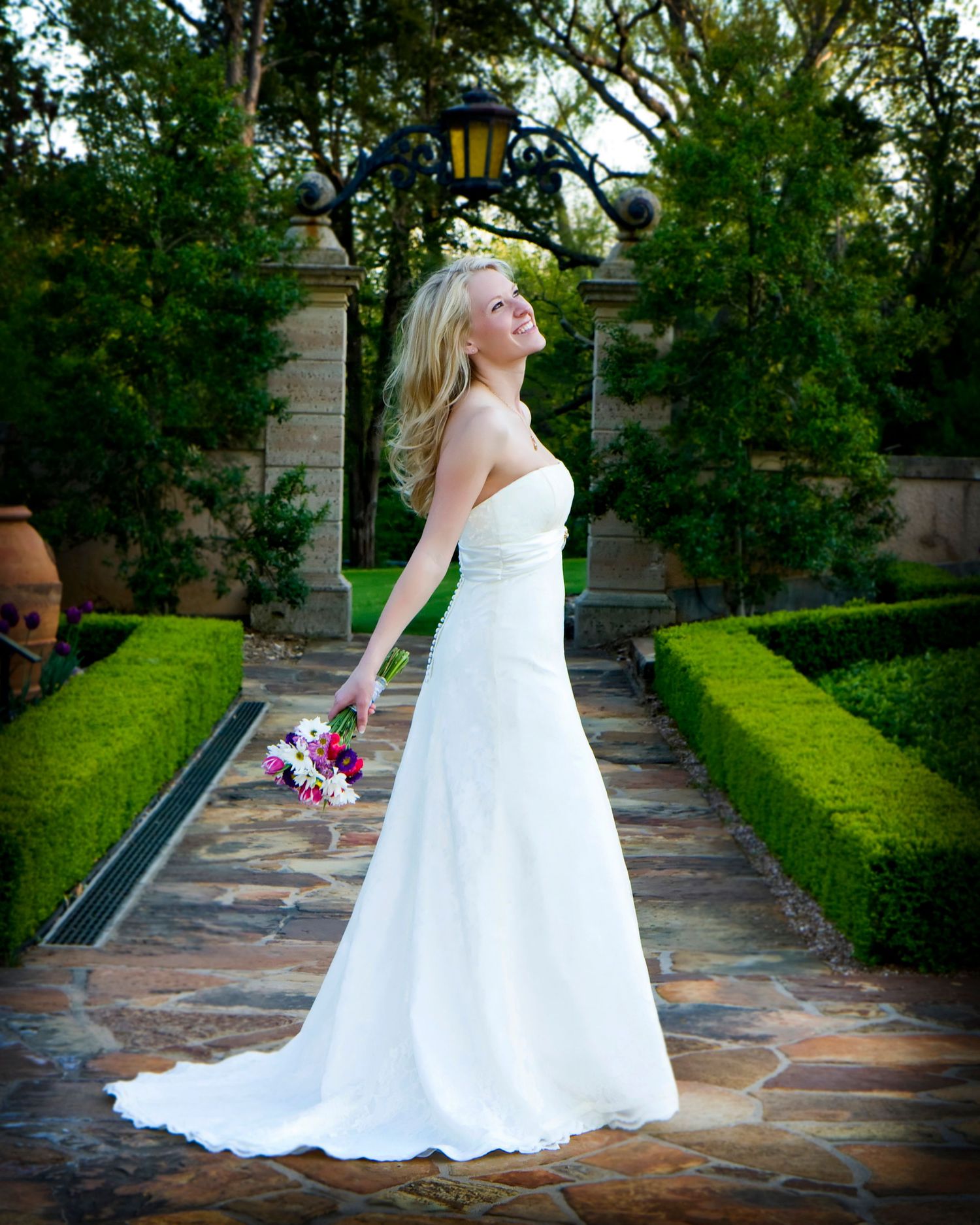 Bride in simple white gown stands on garden path with manicured hedges.