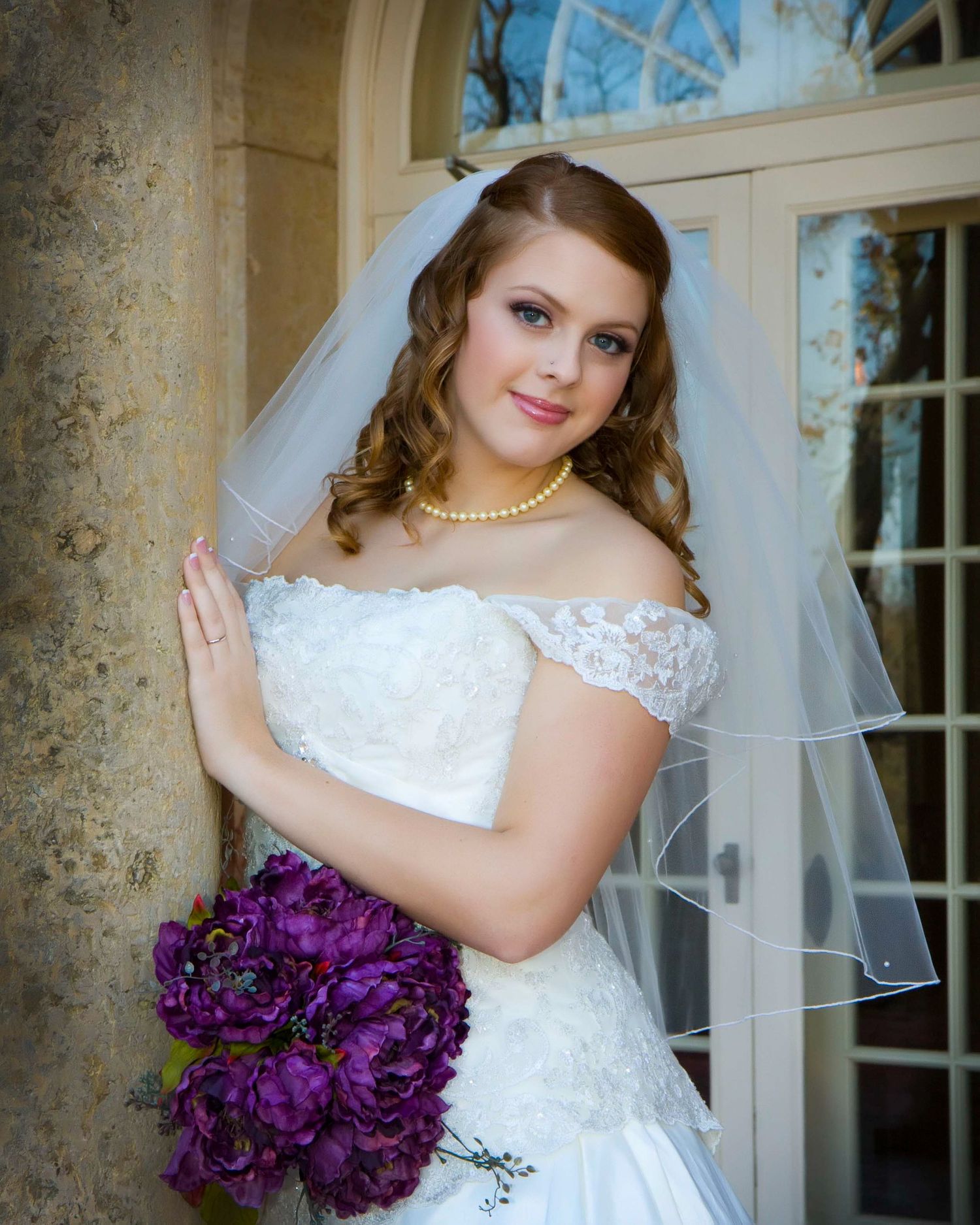 Bride in lace off-shoulder gown holds purple bouquet against stone column.