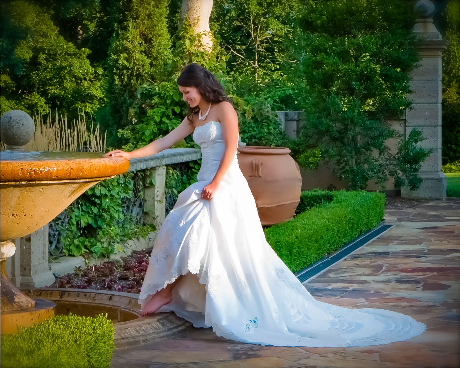 Bride in strapless gown leans on garden furniture amid lush greenery.