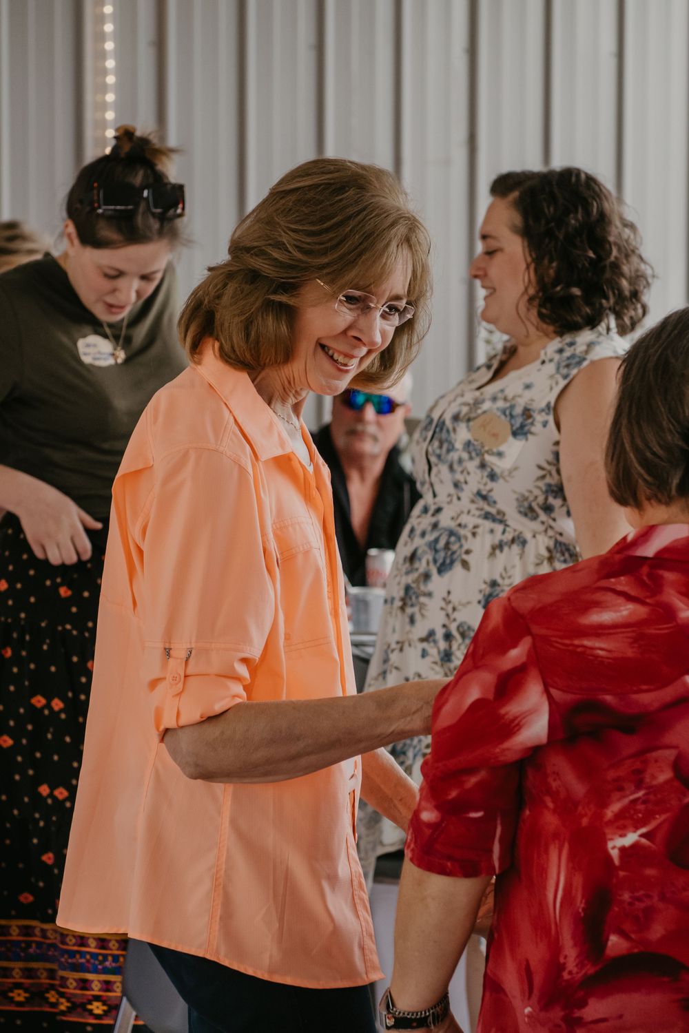 A smiling woman in a peach shirt socializes with guests at an indoor gathering.