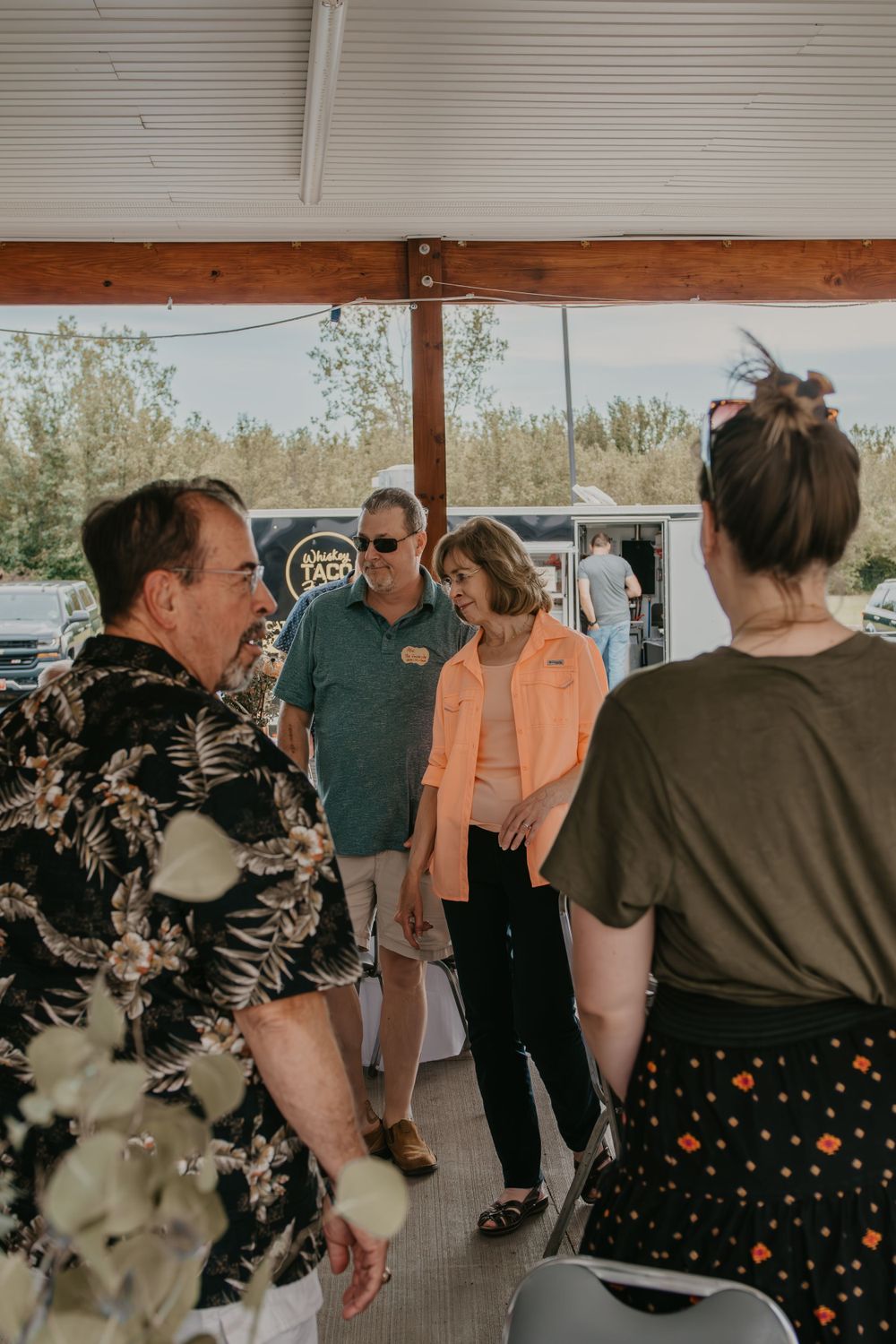 Group of adults in casual attire conversing under an outdoor pavilion at a sunny event.