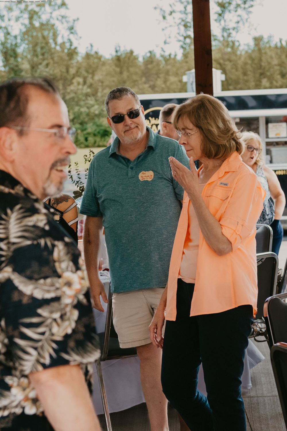 Woman in peach shirt gestures while talking to a man in a floral shirt at an outdoor event.