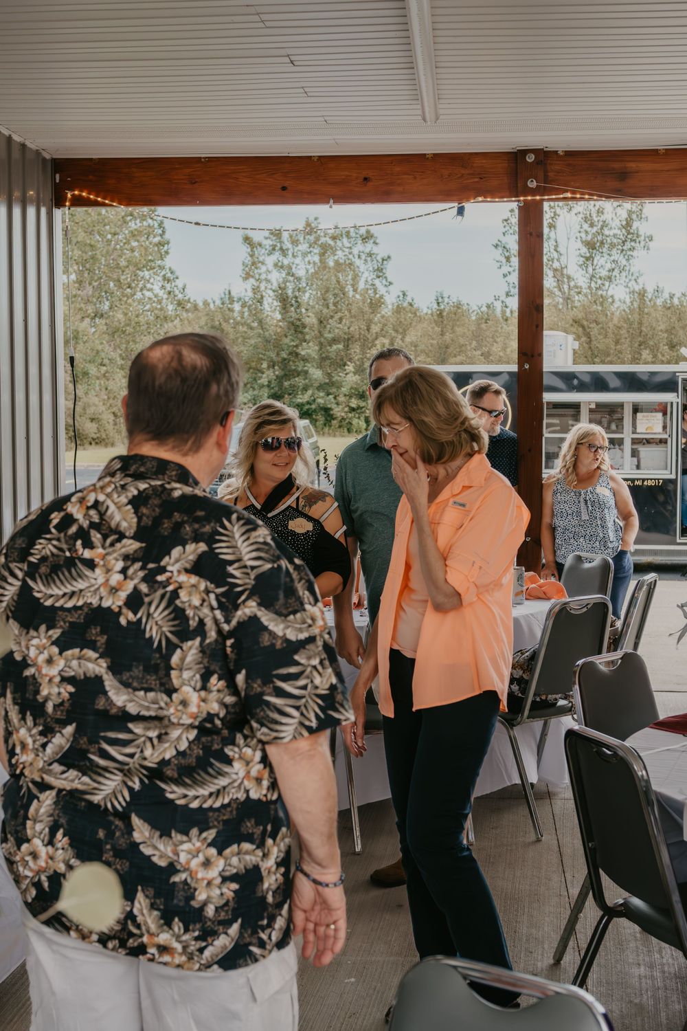 Adults mingling under a wooden pavilion at an outdoor daytime social gathering.