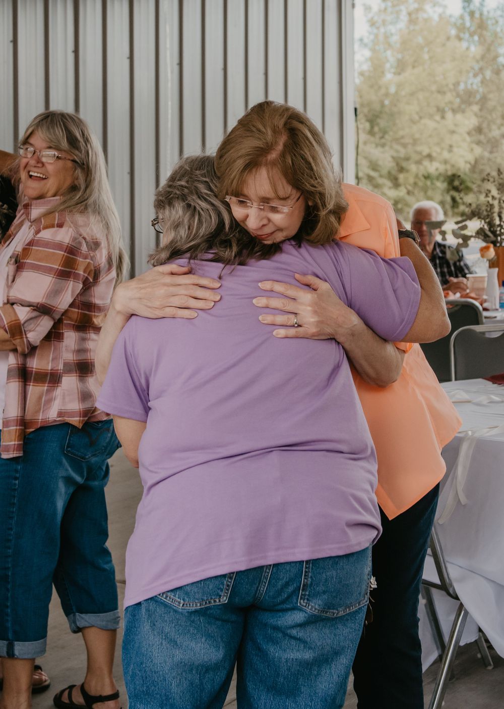 Two women share a warm embrace at a casual outdoor gathering in a barn venue.