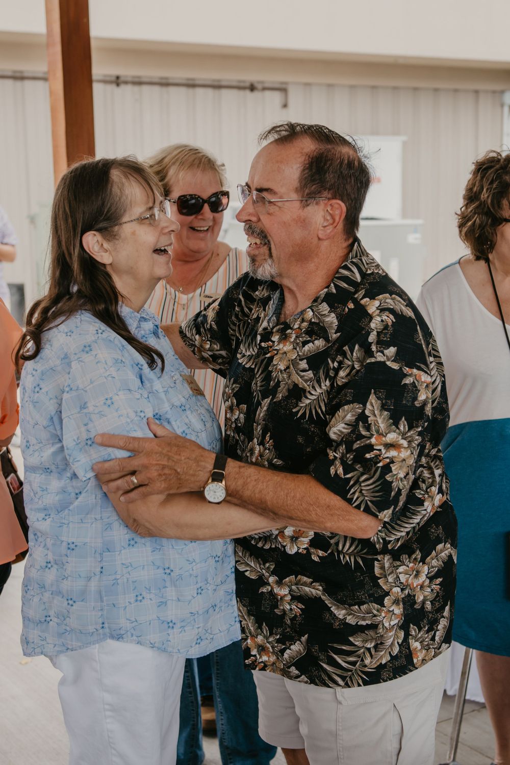A man in a floral shirt and a woman in blue share a warm moment at a social gathering.