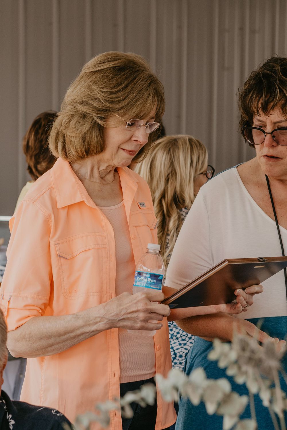 A woman in a peach shirt holds a water bottle while reviewing a framed document at an event.