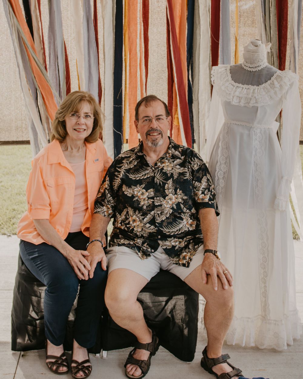 Couple posing together in front of colorful ribbon backdrop with vintage white wedding dress on mannequin.