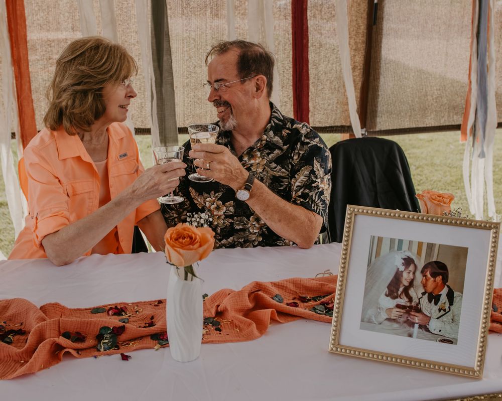 Couple toasting champagne glasses at anniversary celebration table with framed wedding photo.