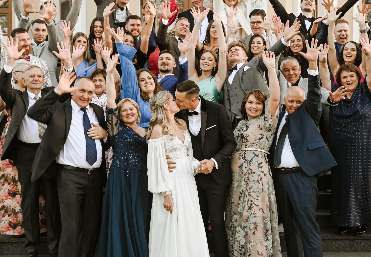 Large wedding party celebrates joyfully with raised arms and excited expressions during outdoor group photo.