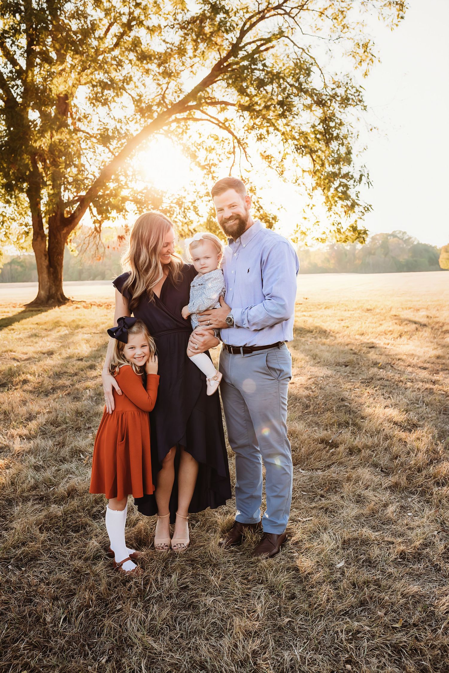 Family portrait at golden hour in field with sunlight streaming through tree branches.