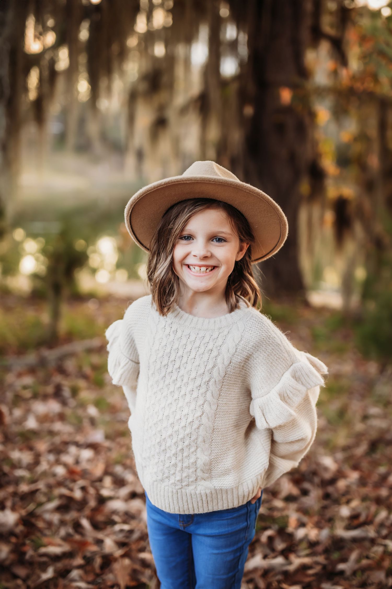 Child wearing cozy cream cable knit sweater and tan hat poses in autumn forest with falling leaves.
