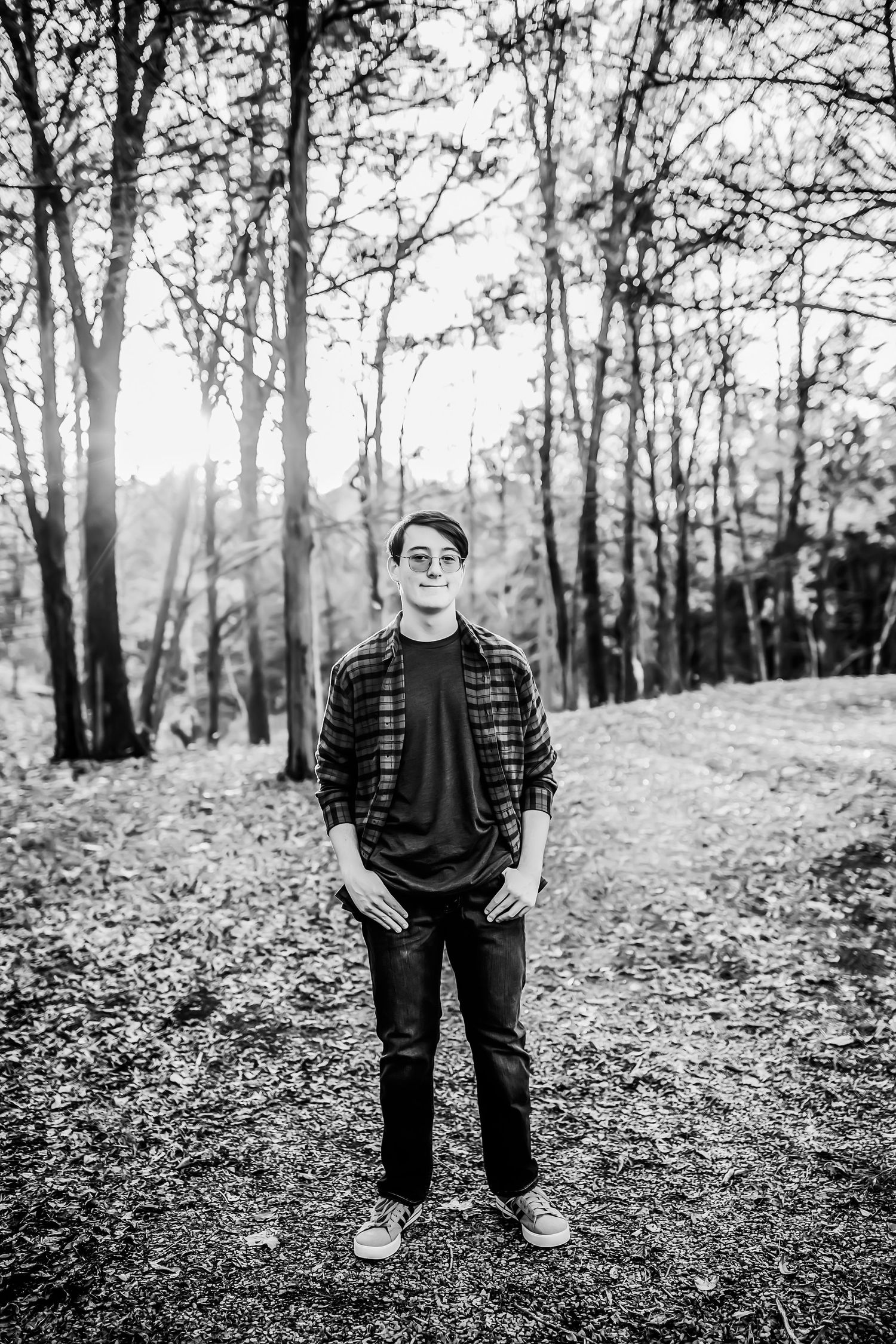 Black and white portrait of someone in casual attire standing on a leaf-covered forest path.