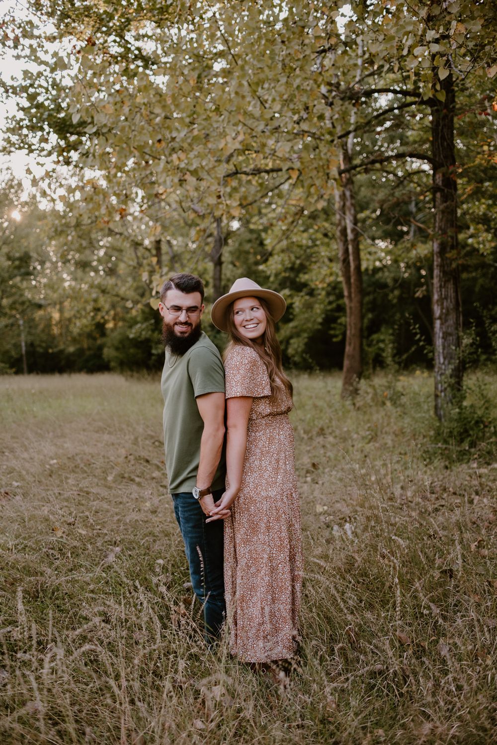 A couple poses romantically among tall grass in a wooded area.