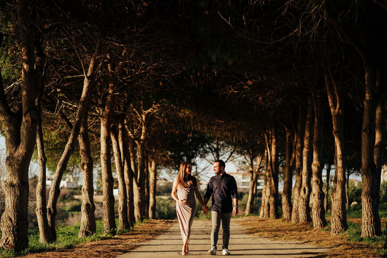 A couple walks hand in hand down a tree-lined path during golden hour sunset.
