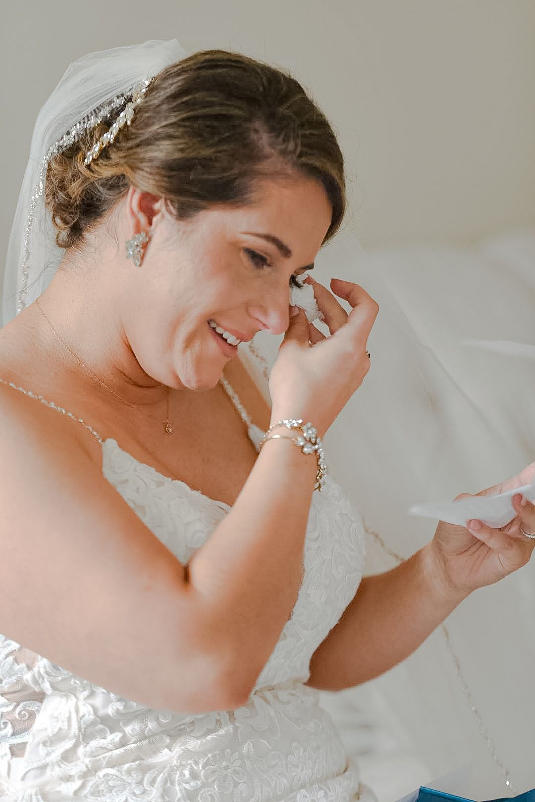 Emotional bride in white dress and veil wiping tears while reading note.