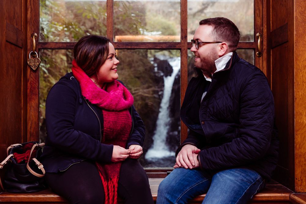 Two people engaged in conversation by a window overlooking a scenic waterfall.