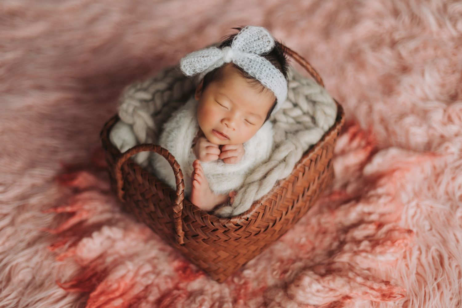 Newborn baby sleeping peacefully in a wicker basket with a gray knit bow headband on a pink fluffy rug.