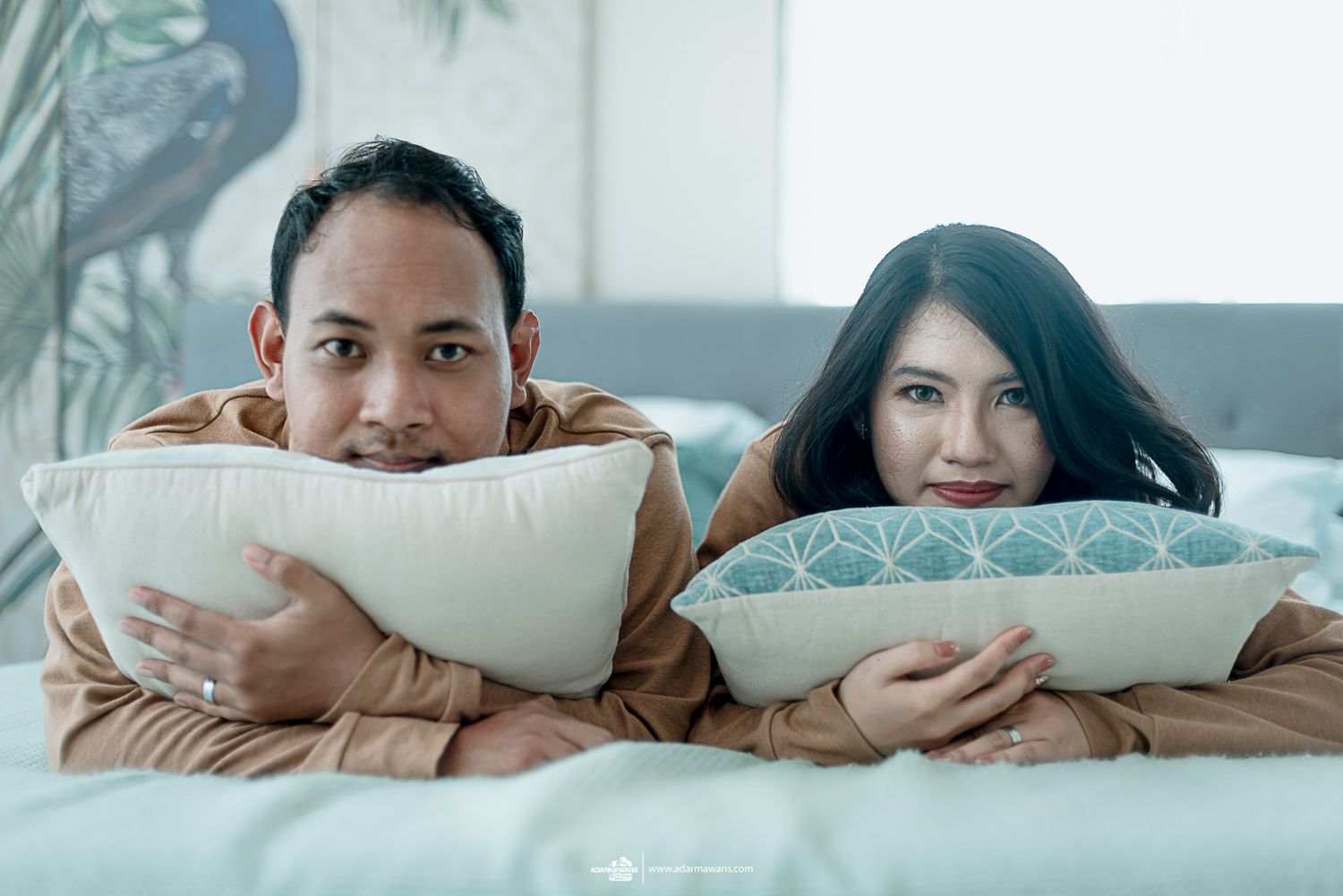 Couple peeking over decorative throw pillows while relaxing on bed together.