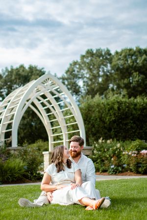 Couple sitting on grass in front of white garden arch on a cloudy summer day.