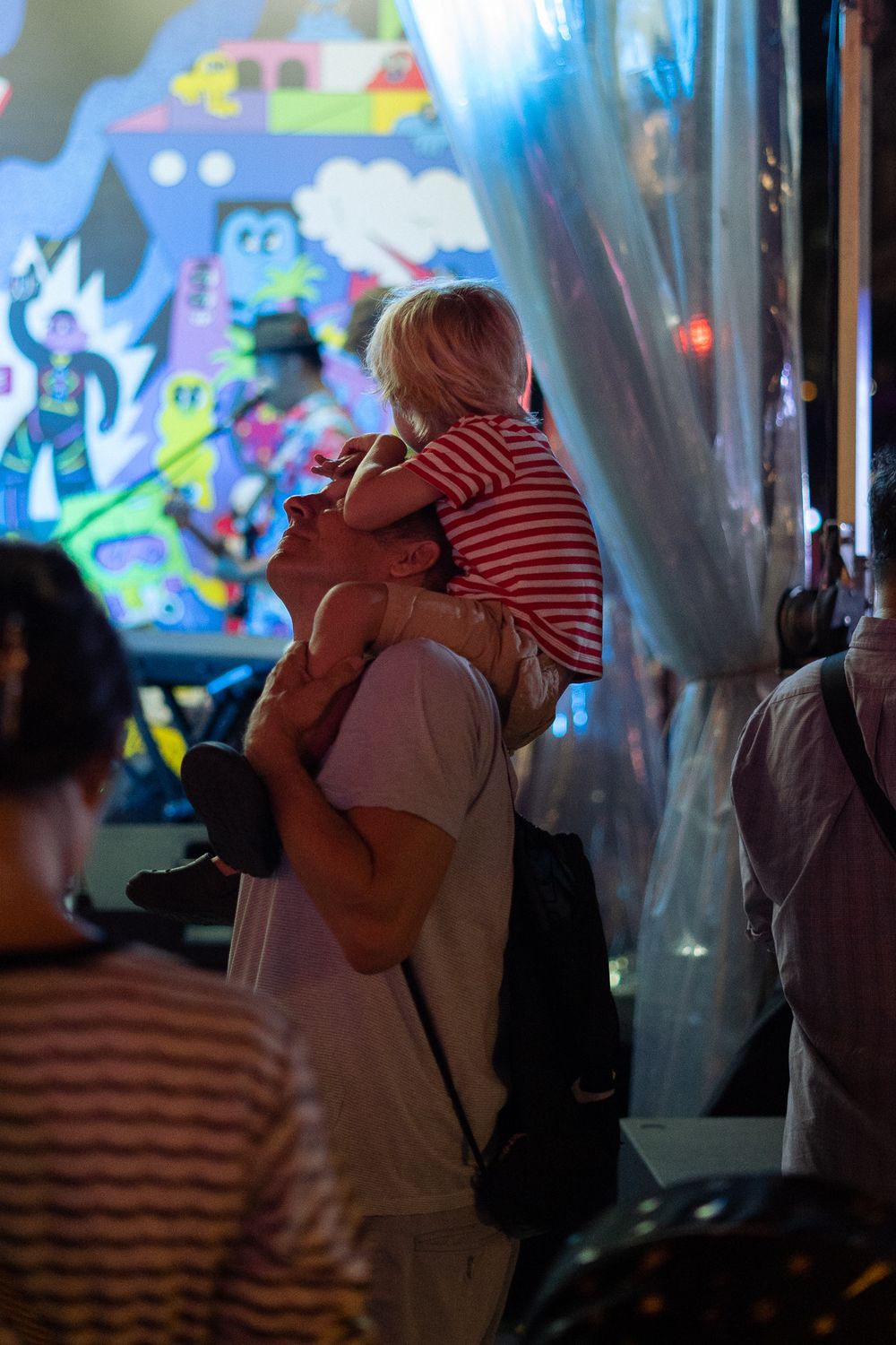 A silhouetted figure carries a child on shoulders against a colorfully lit backdrop at night.