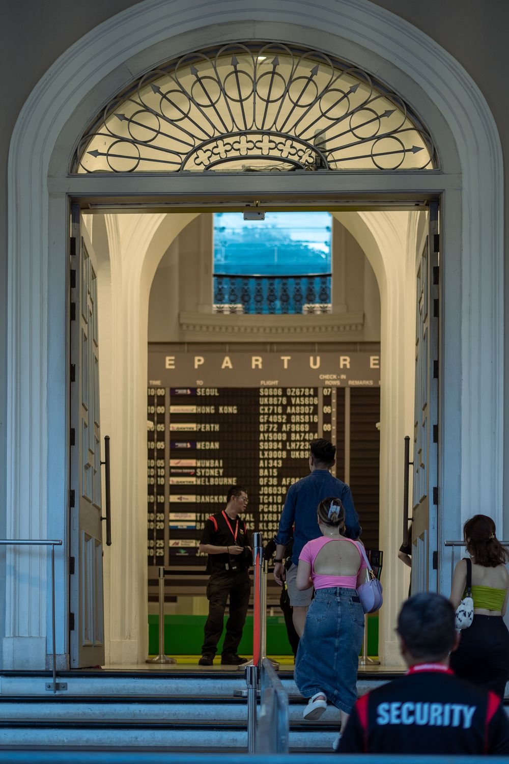 People queue at an ornate arched entrance with a departure board display.