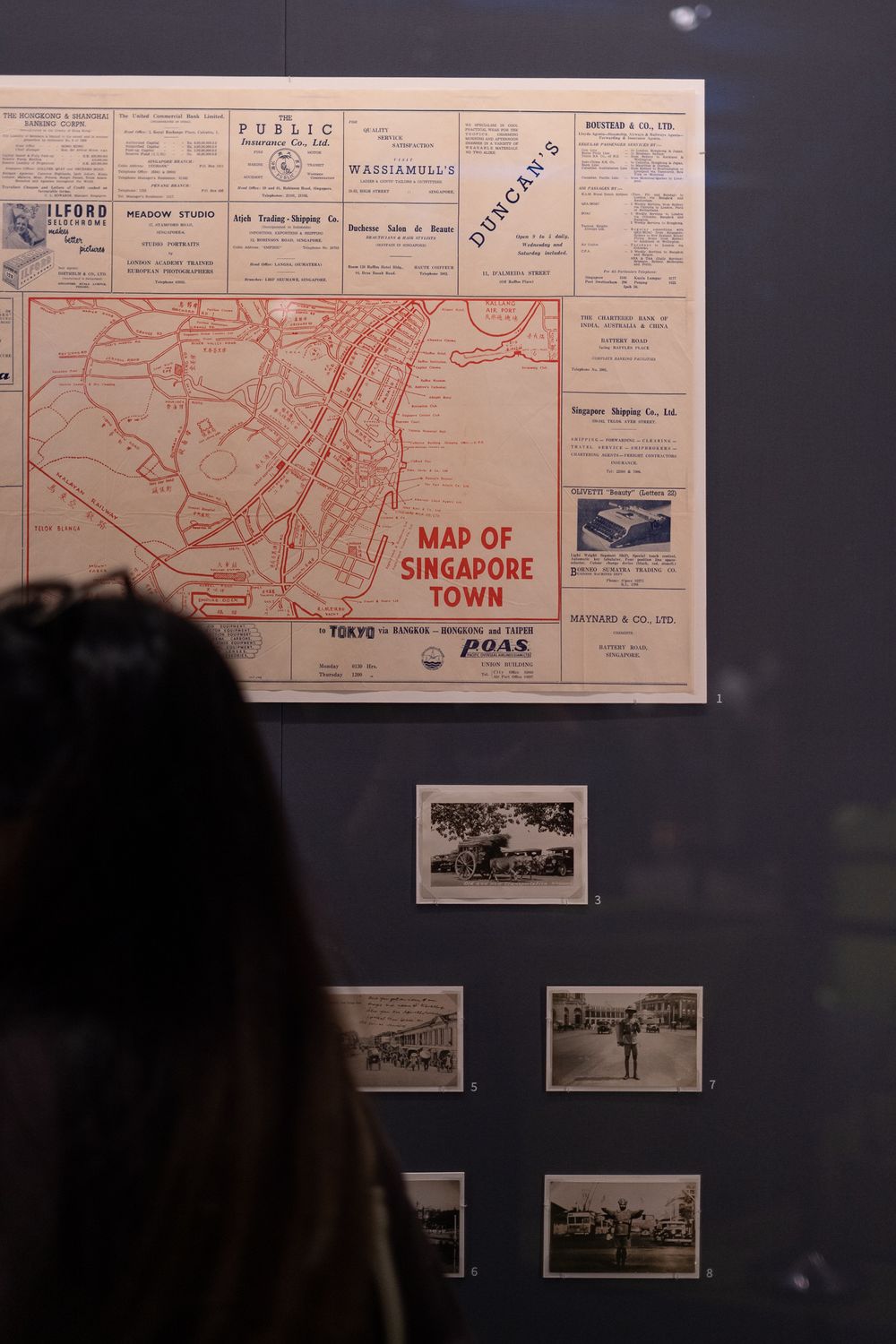 Vintage red map of Singapore Town displayed on wall with black and white historical photographs below.