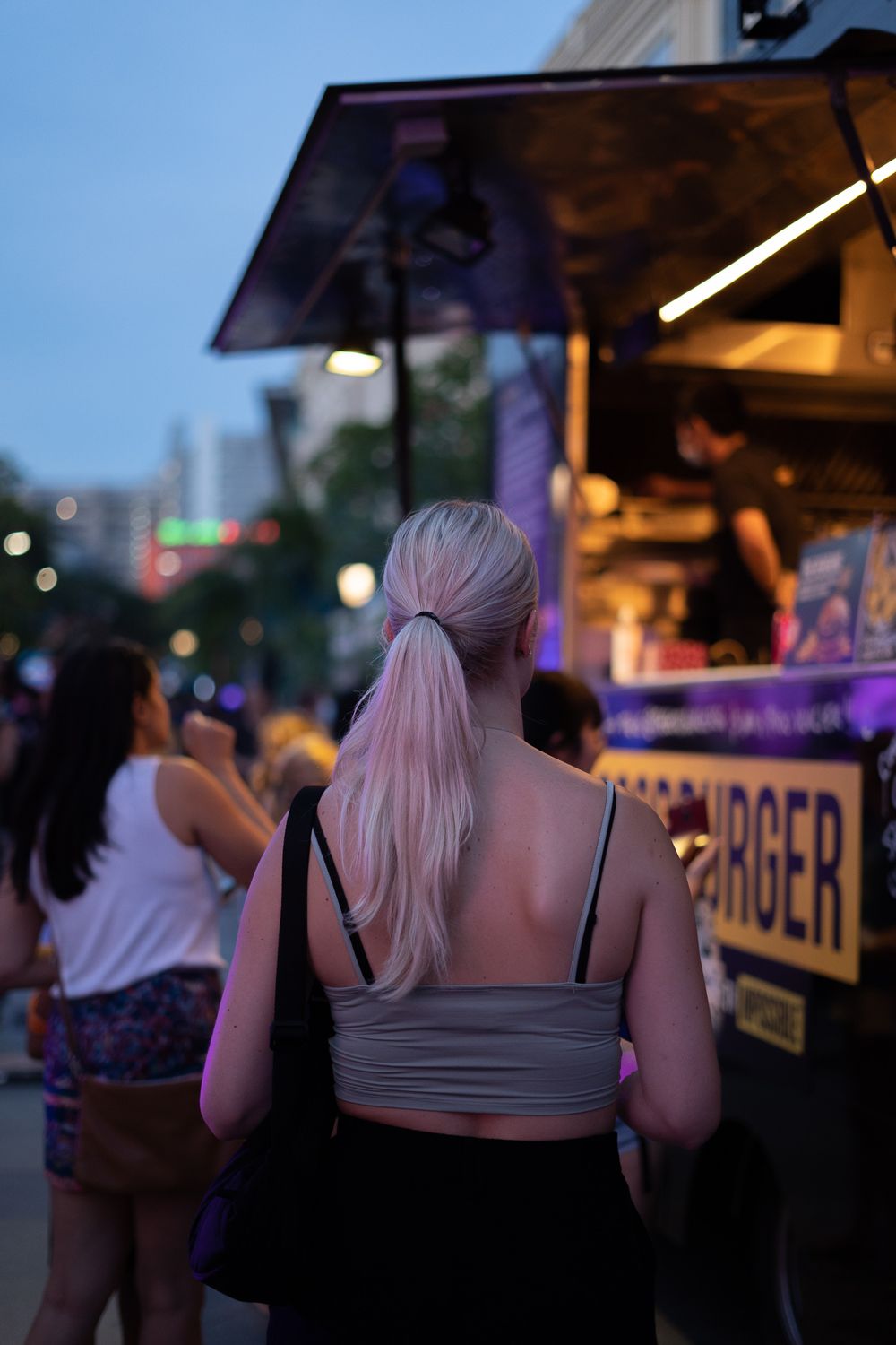 People gather at a food vendor stand in the evening with illuminated city lights in the background.