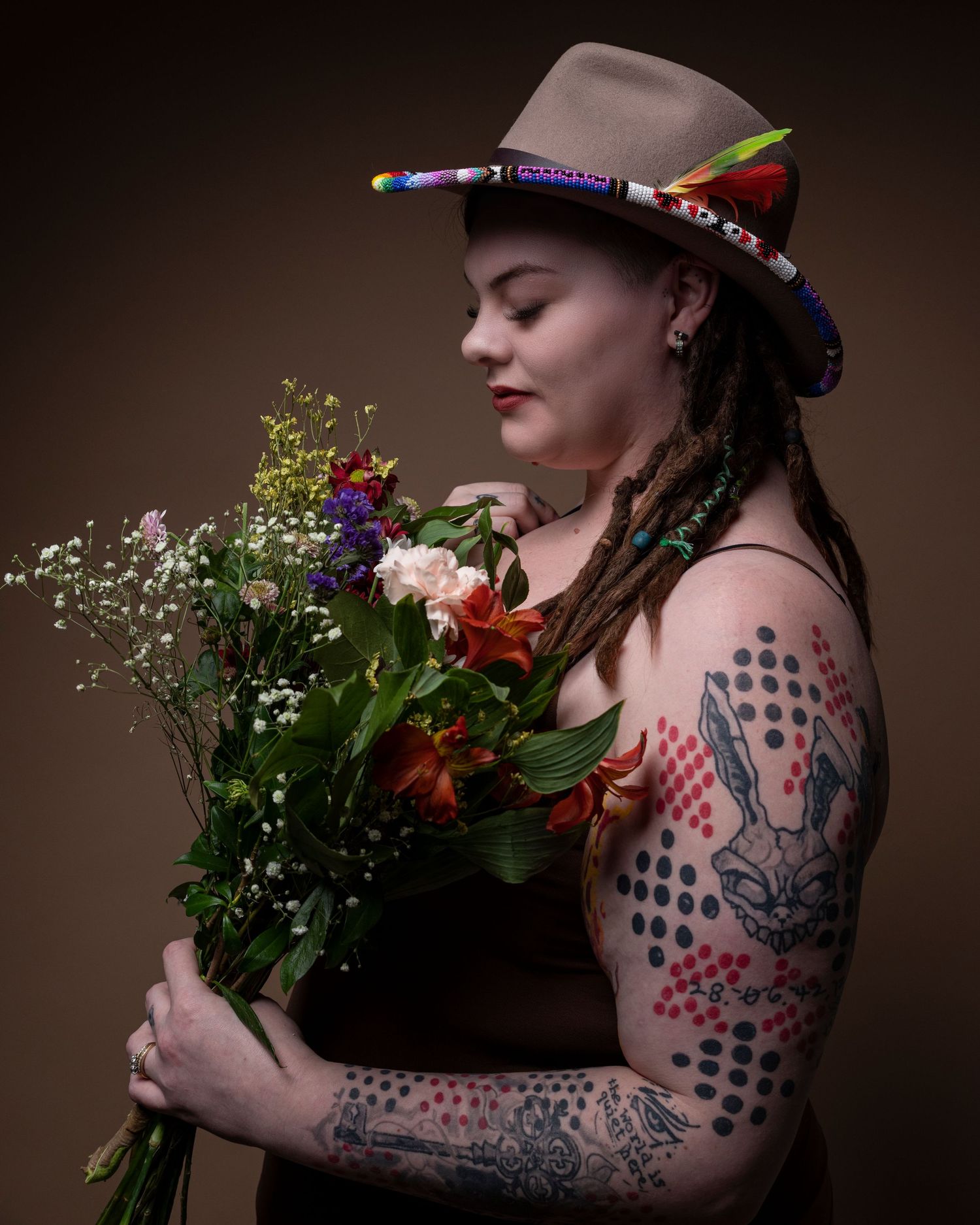 Portrait of someone holding a vibrant bouquet while wearing a hat and displaying ornate arm tattoos against a brown backdrop.