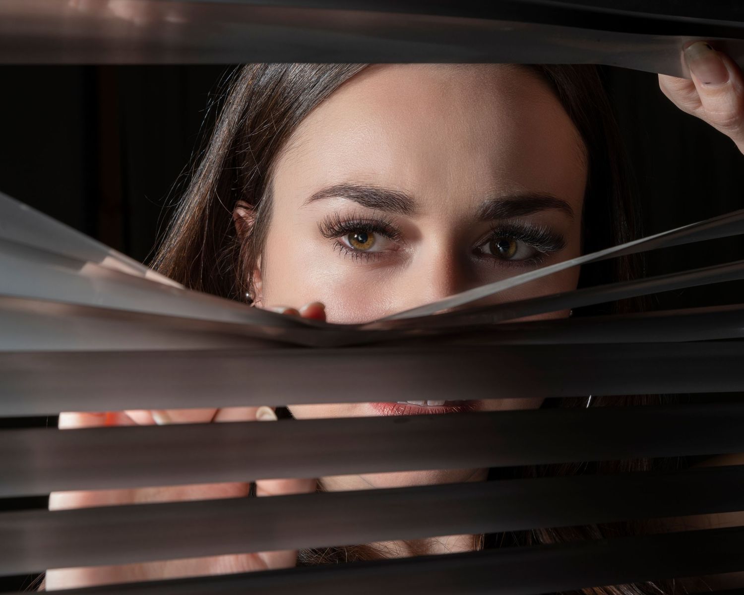 Artistic portrait peering through window blinds with dramatic lighting.