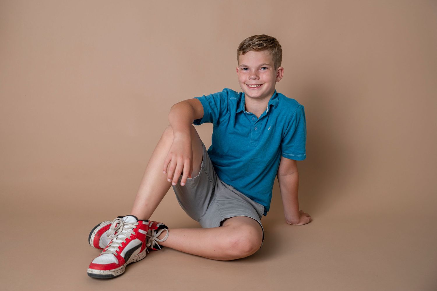 Youth in blue polo shirt and grey shorts sitting casually against beige background.