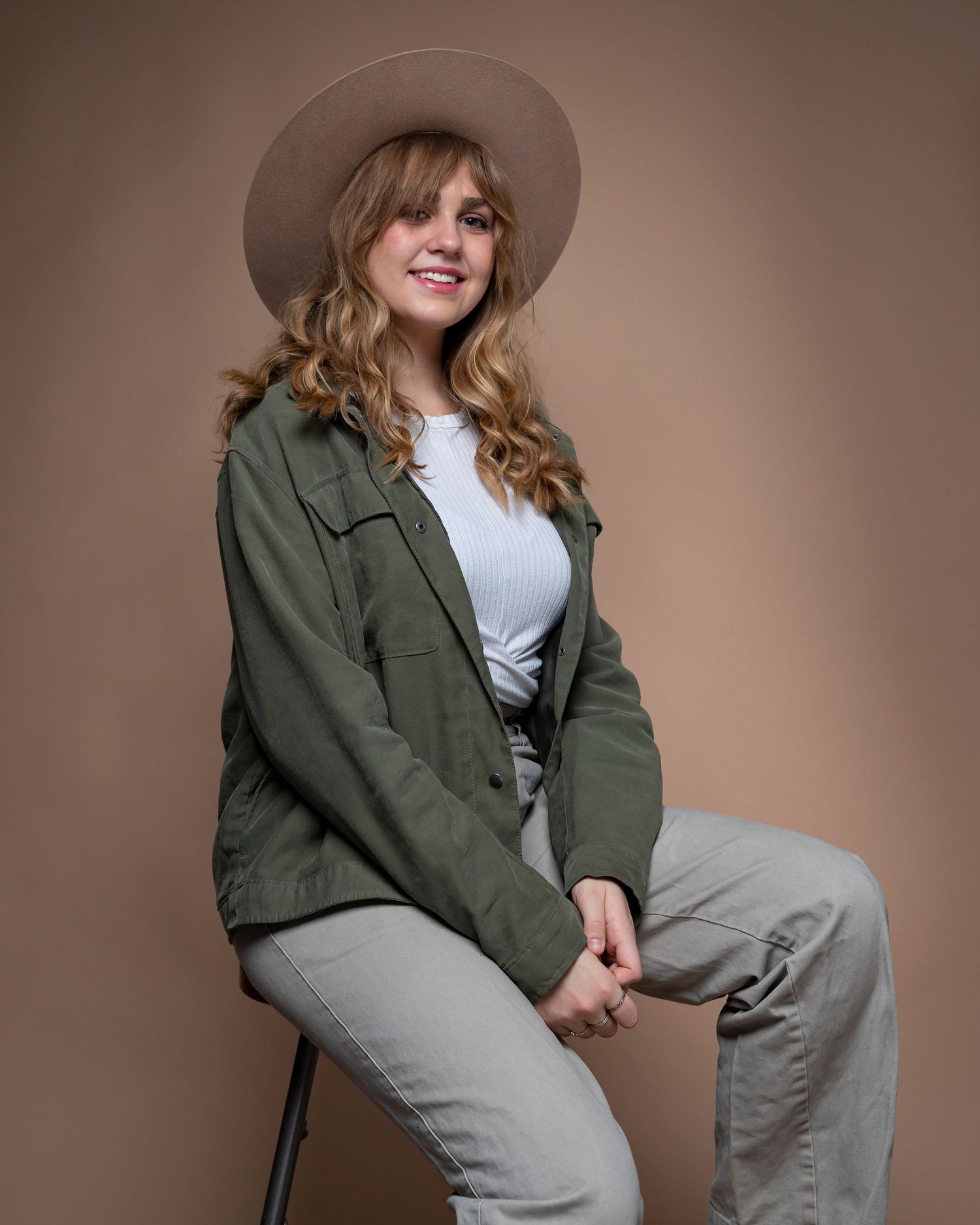 Fashion portrait featuring someone in a tan hat, olive jacket and gray pants seated on a stool.