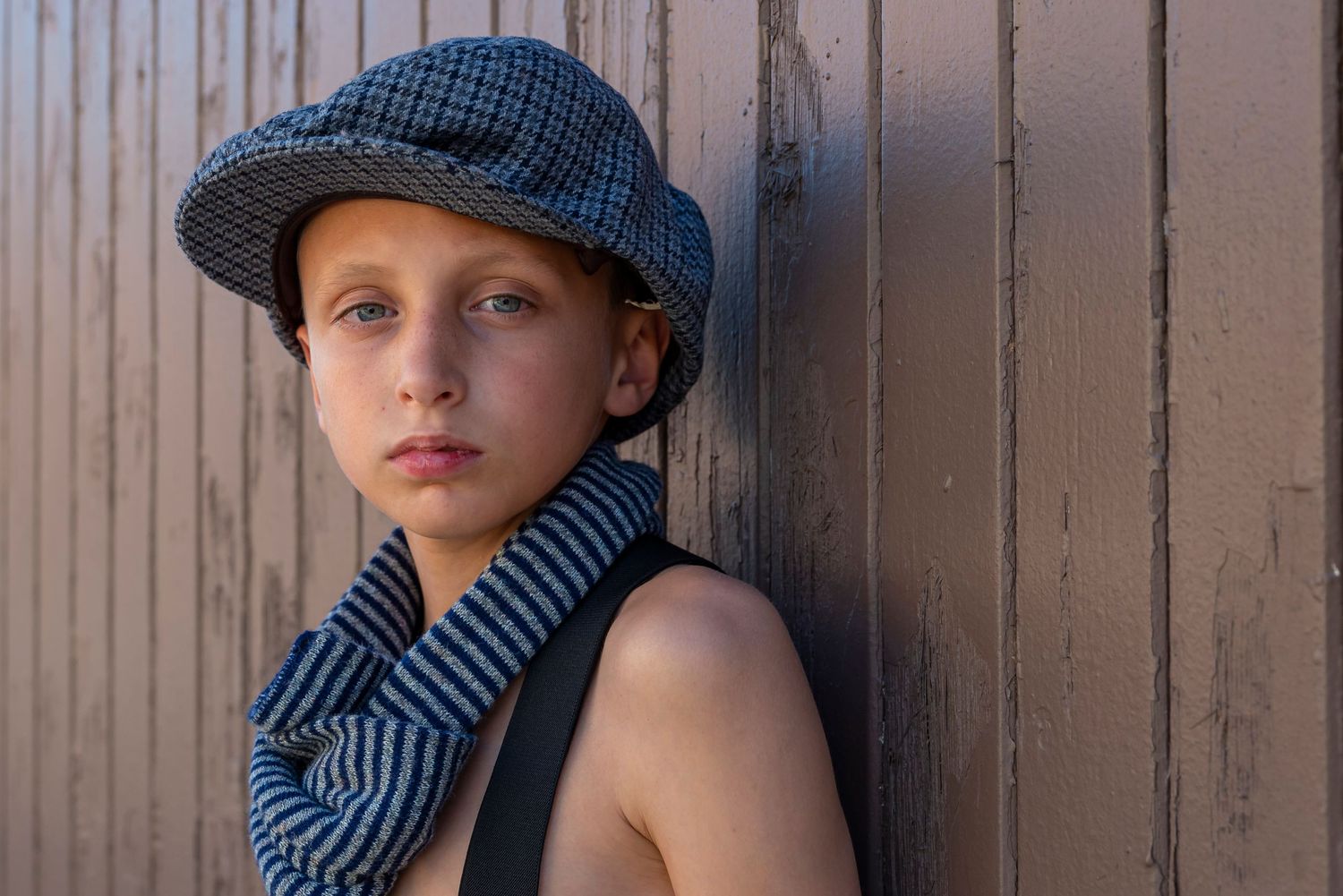 Portrait wearing newsboy cap and striped scarf against wooden wall.