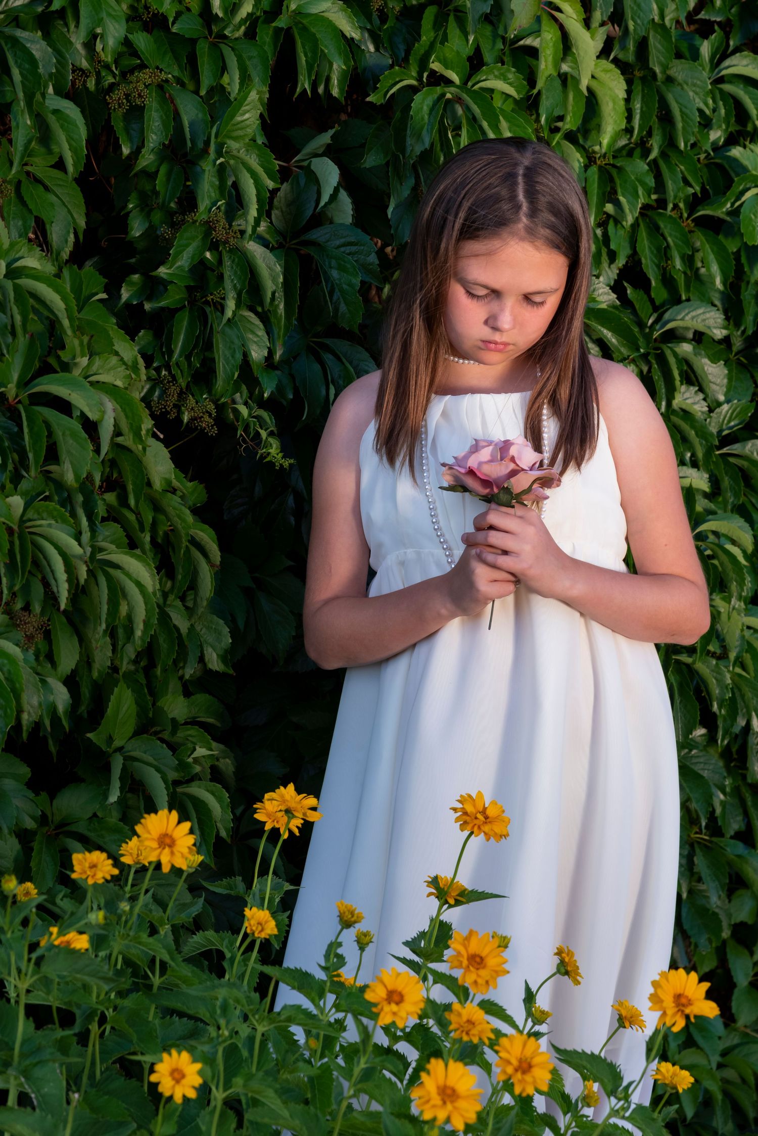 Young woman in white dress holding flowers stands against green foliage with yellow daisies blooming below.