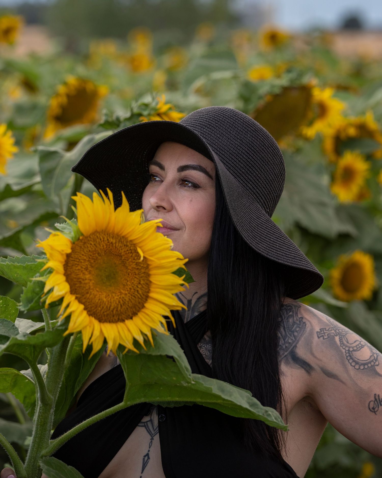 Woman in black hat posing next to large sunflower in field.