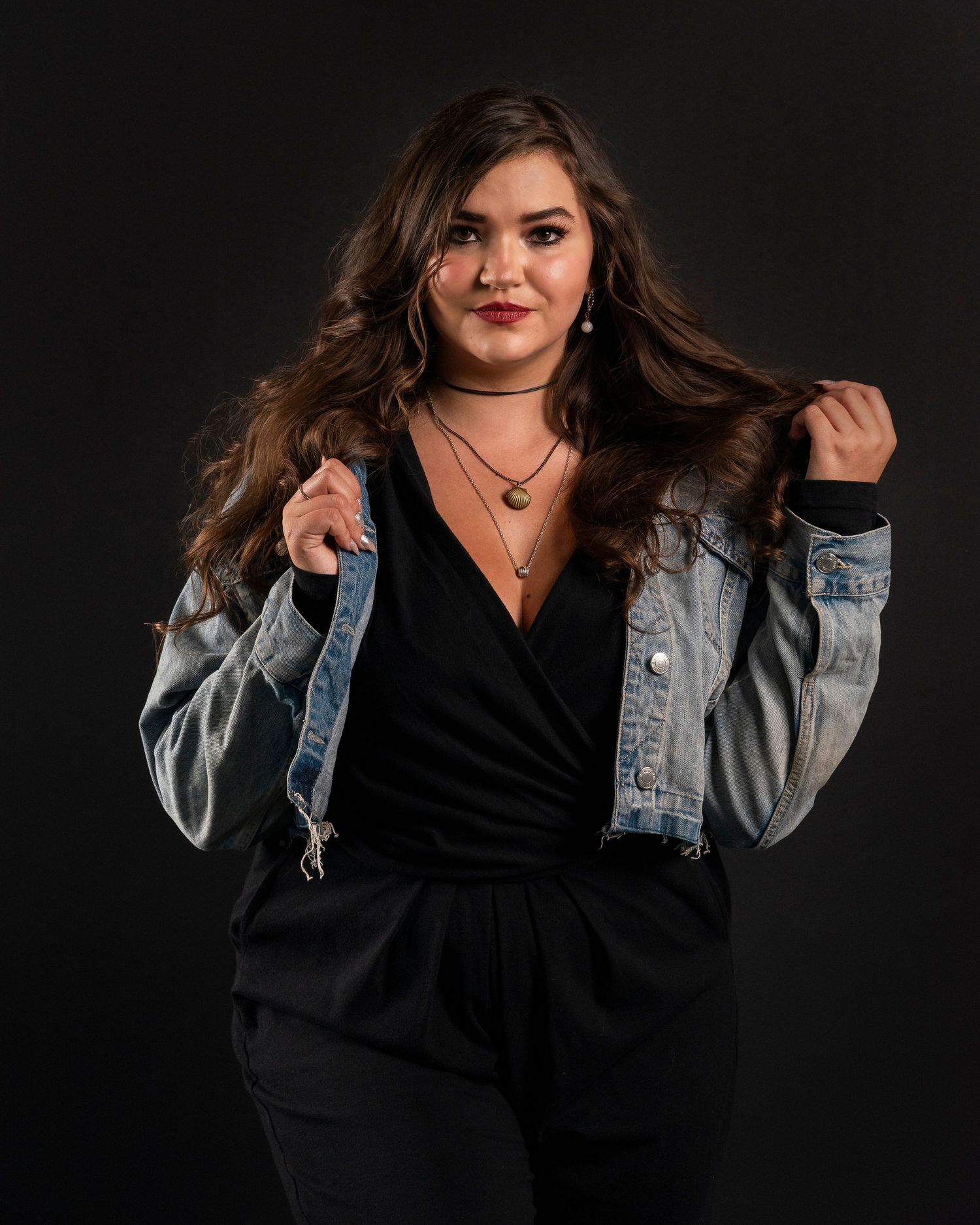 Fashion portrait with someone wearing a denim jacket and black outfit against dark background.