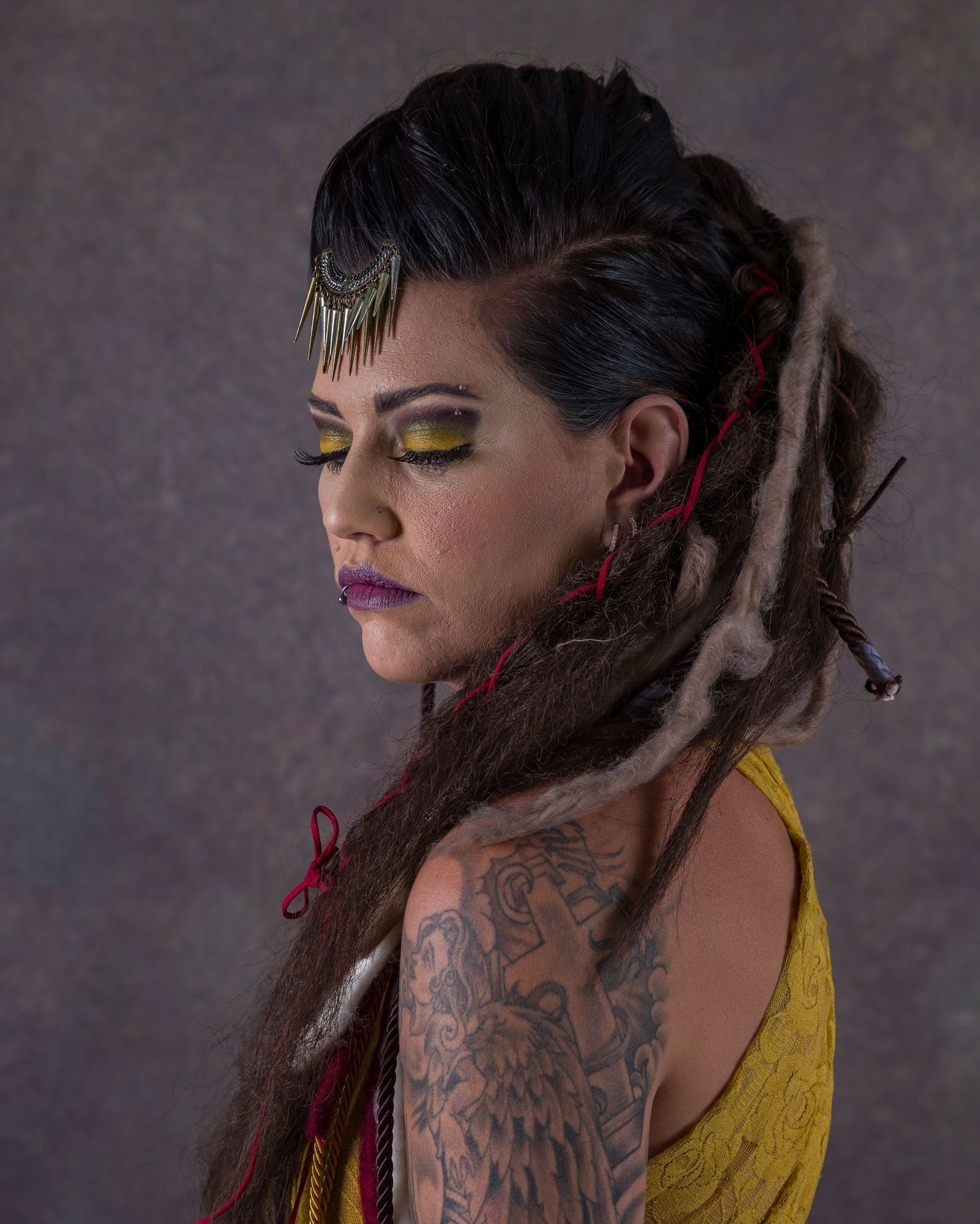 Creative portrait showing dramatic makeup and dreadlocks with yellow clothing against purple backdrop.