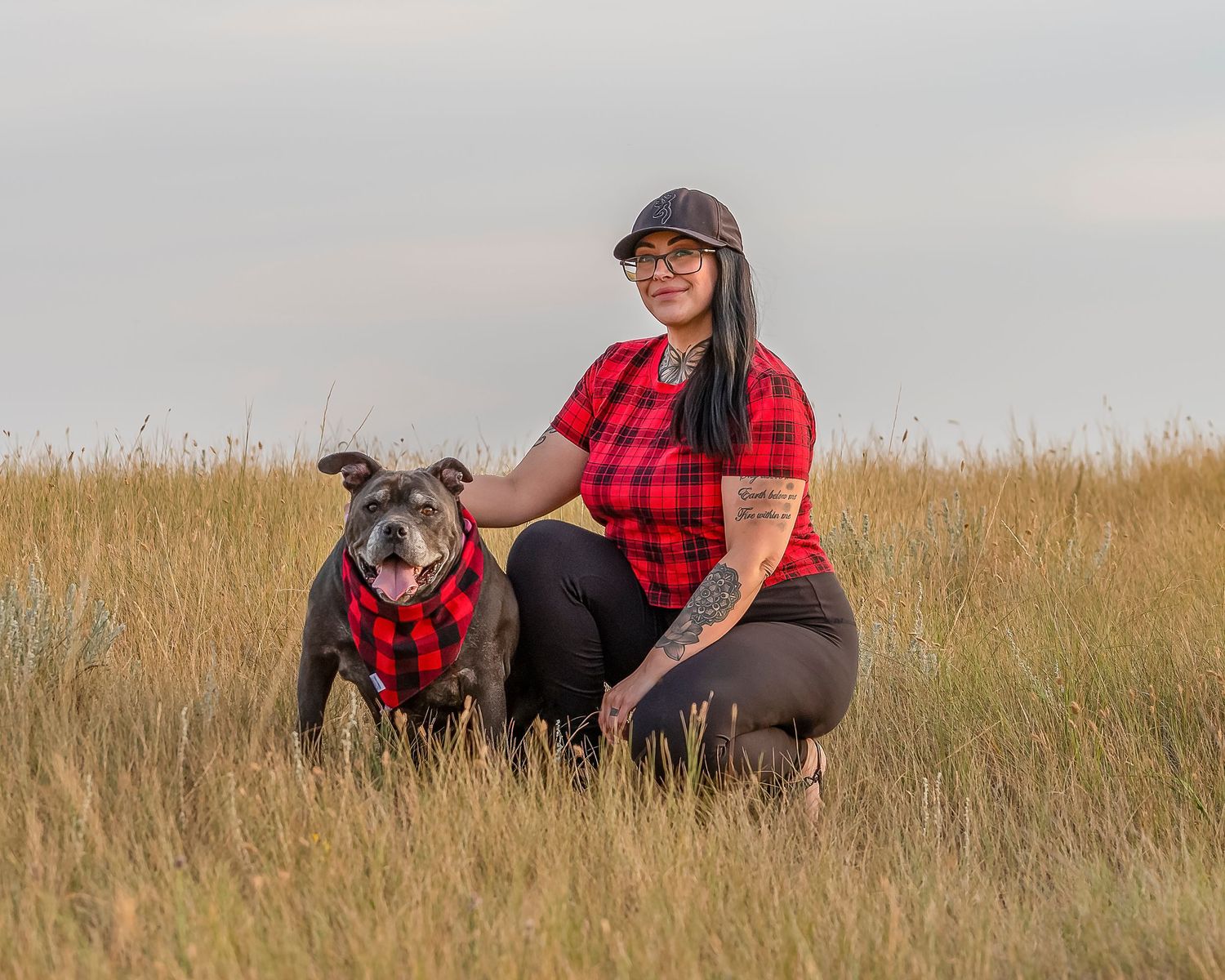 Person and dog wearing matching red plaid shirts sitting in tall grass at sunset.