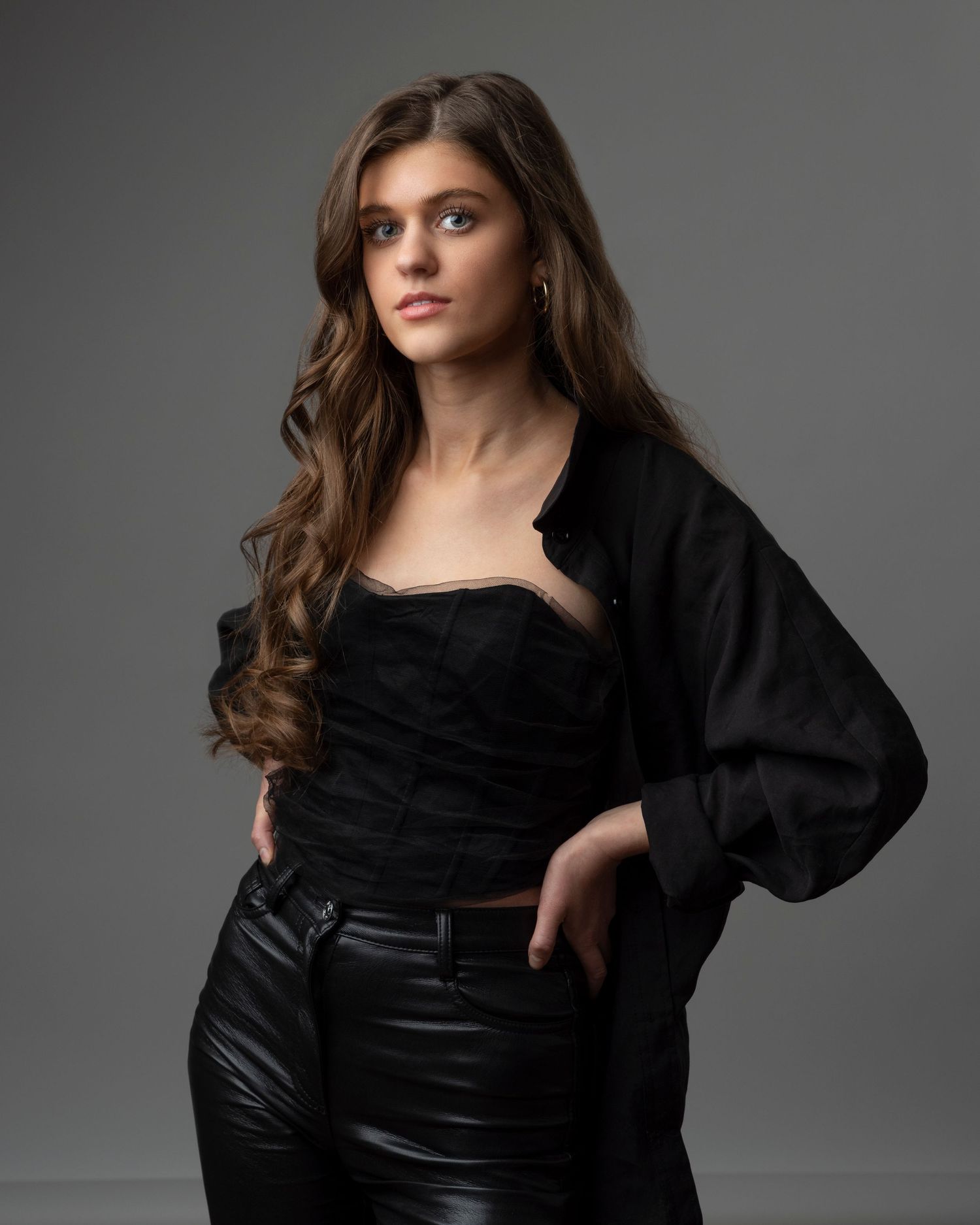 Fashion portrait in black outfit with flowing dark hair against grey backdrop.