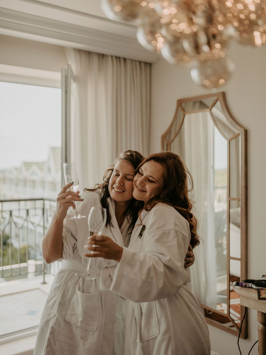 People in white robes sharing champagne and laughter in an elegant room with chandelier lighting.
