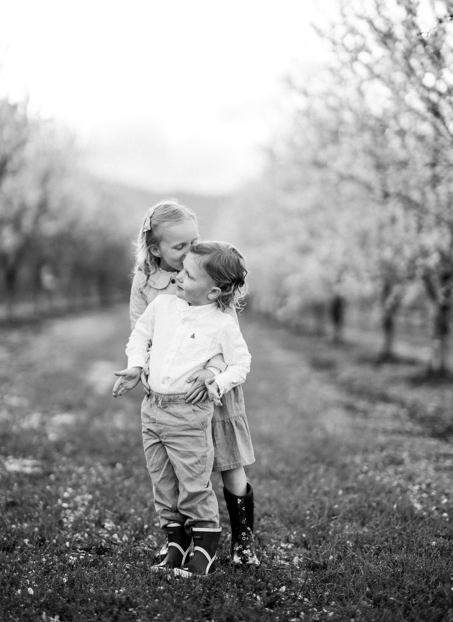 Two children share a tender moment in an orchard with bare trees in black and white.