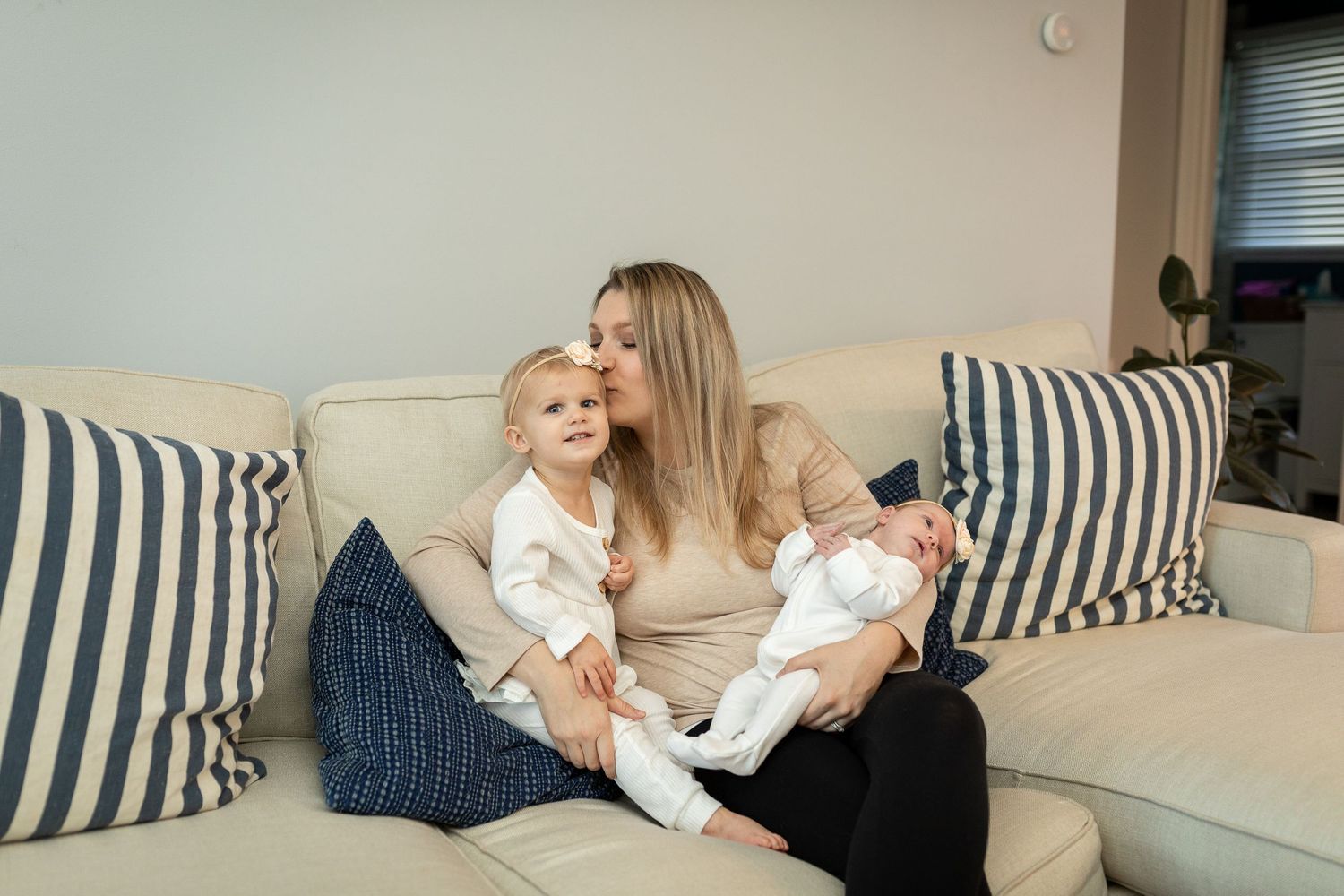 Parent sits on couch with toddler and baby in white outfits.