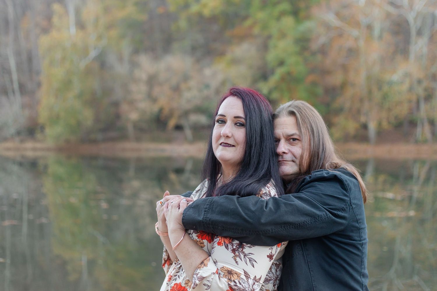 Two people embrace while standing by a tranquil lake during autumn.
