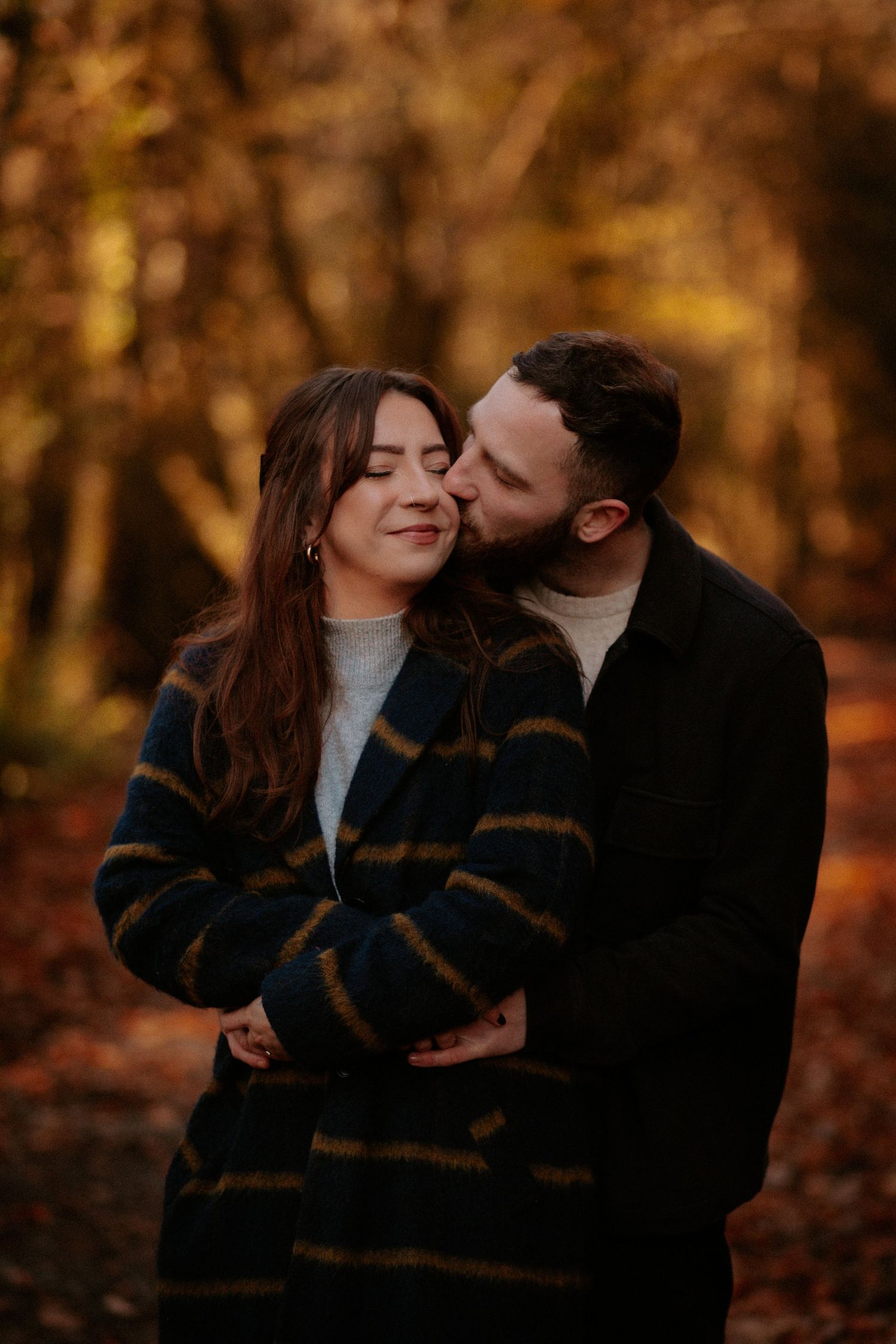 Intimate couple shares tender moment against autumn forest backdrop wearing cozy winter coats.