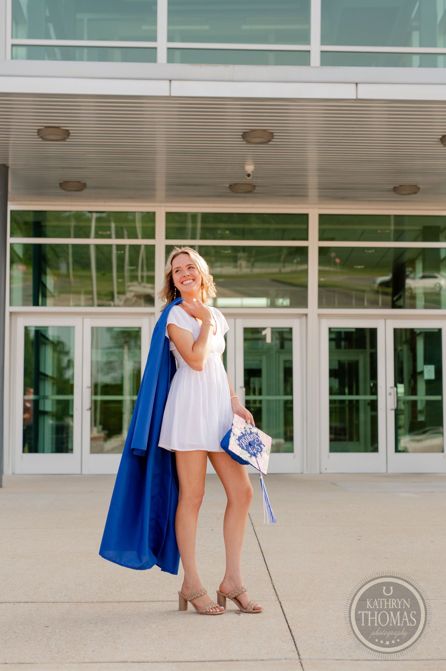 Graduate in white dress and blue gown poses joyfully outside modern school entrance.
