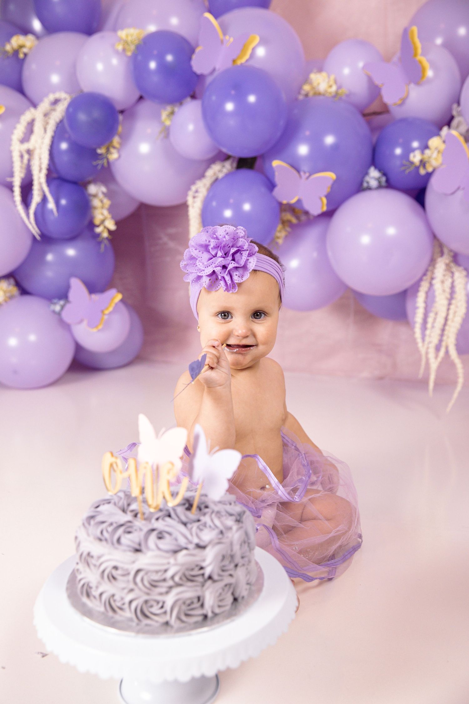 A baby sitting in front of a cake with purple balloons.