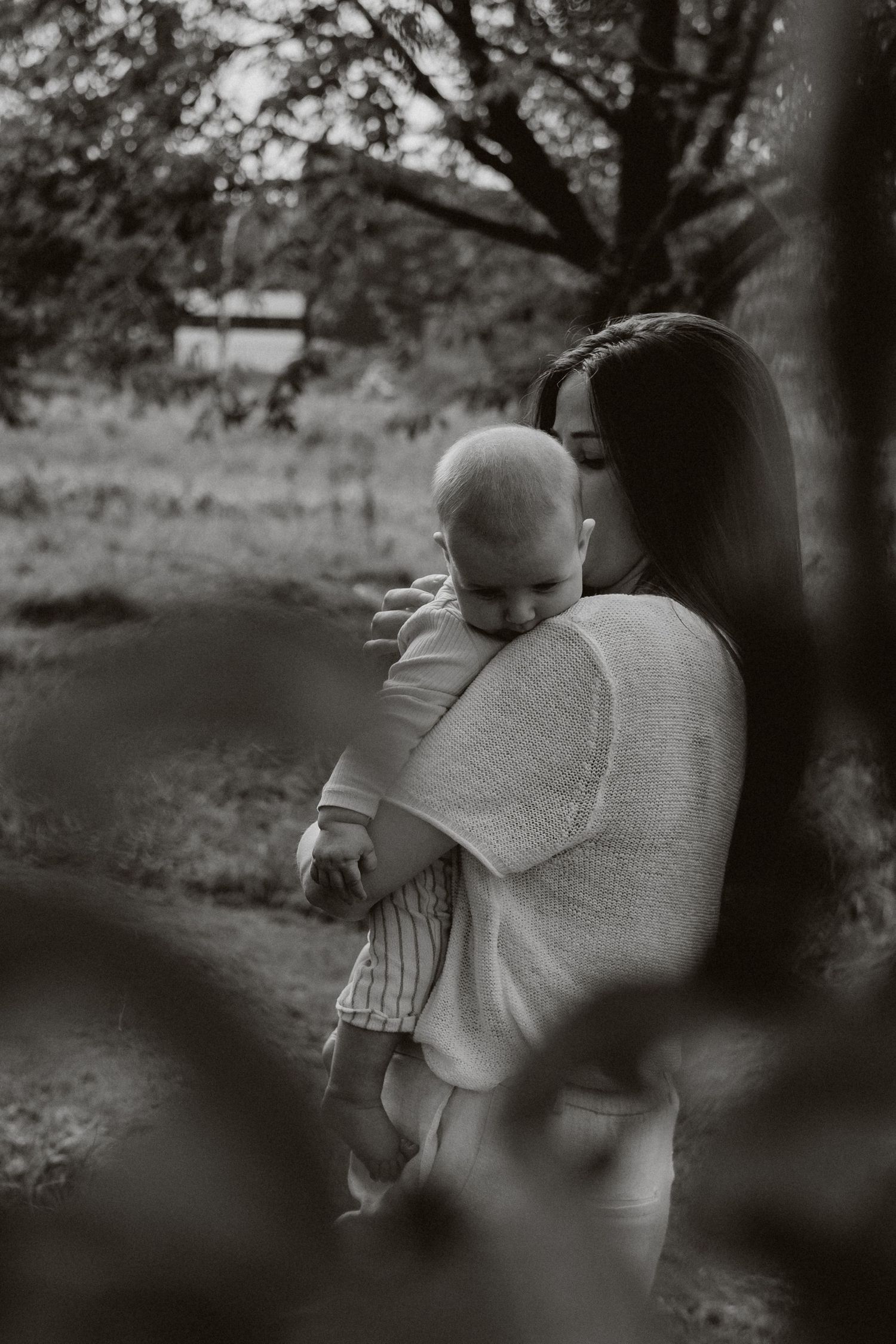 A mother tenderly holds her baby in a black and white outdoor portrait beneath a tree.