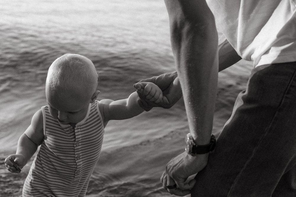 Black and white photo of a small child in striped shirt holding hands with an adult near water.