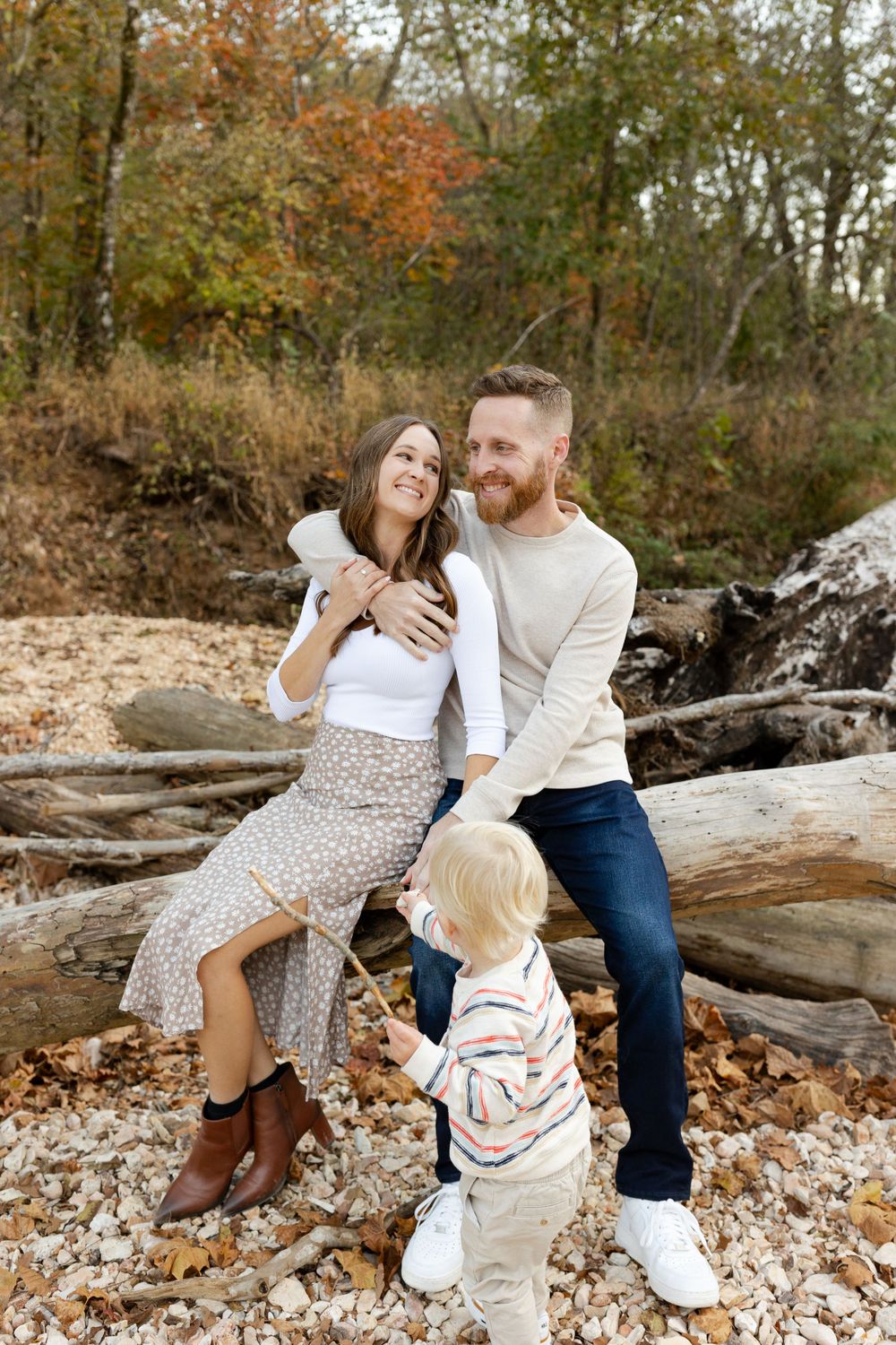 Family sitting together on fallen logs surrounded by autumn foliage in a rustic outdoor setting.