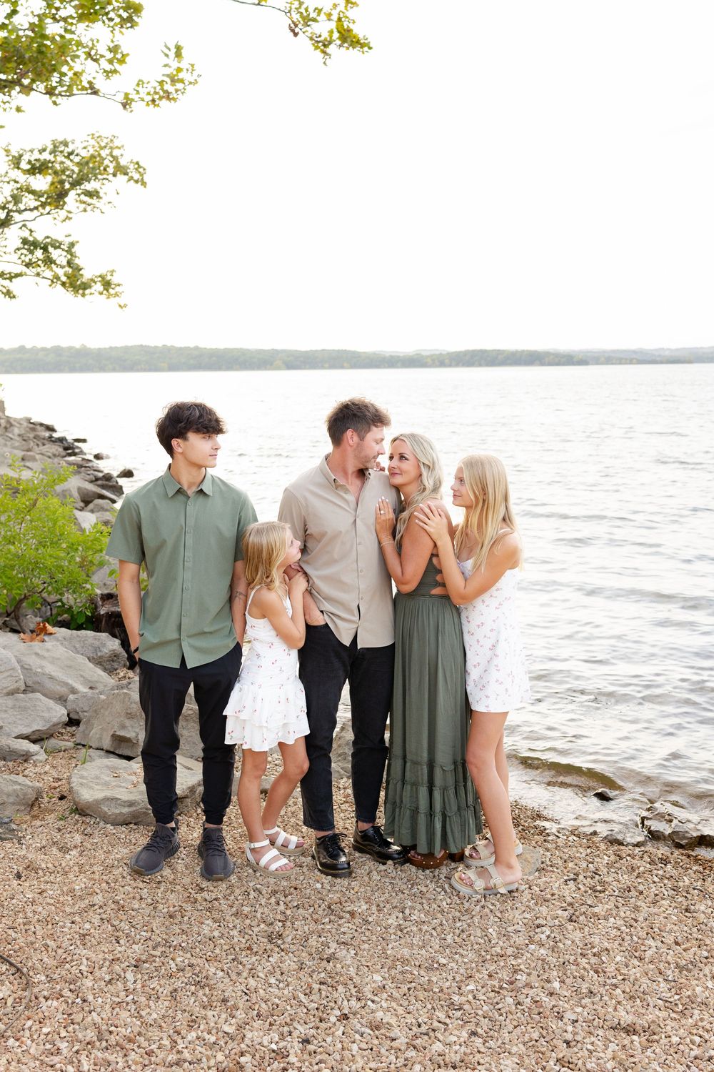 Family group poses together on rocky beach shoreline at sunset with water and trees in background.